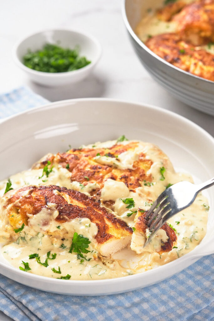 A plate with cooked chicken breast topped with creamy sauce and garnished with chopped parsley.