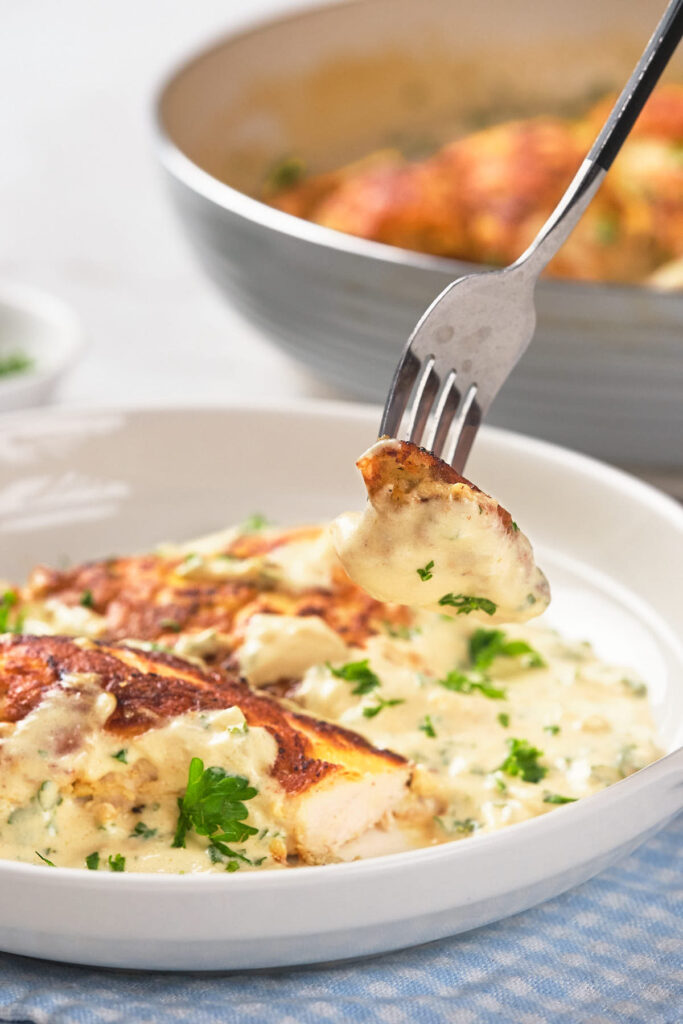 A fork lifting a piece of seasoned chicken breast with creamy sauce from a white plate, garnished with chopped parsley.