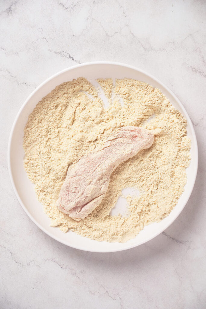 A piece of raw chicken coated in flour lies on a white plate against a light background.