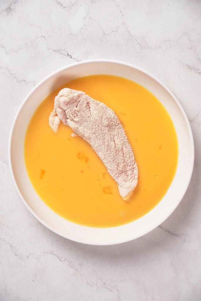 A piece of raw chicken coated in flour is placed in a white bowl of egg wash, on a marble countertop.