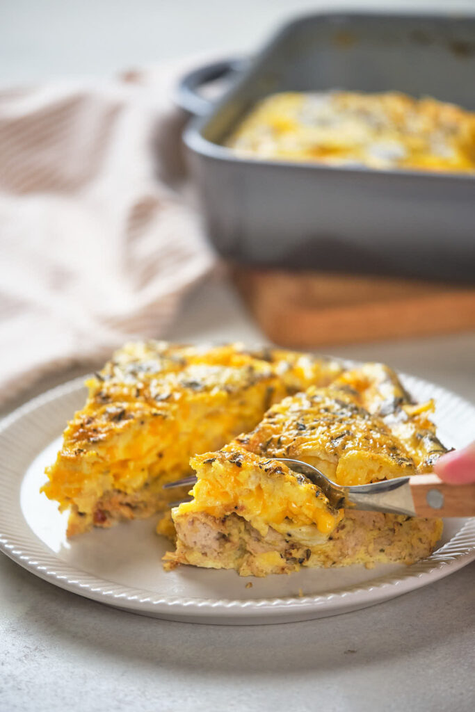 A person cutting a slice of cheesy casserole on a white plate with a fork.