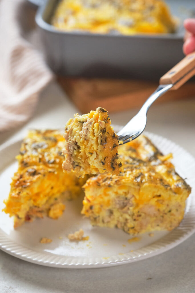 Close-up of a fork holding a bite of baked casserole, with two slices of the casserole on a white plate.