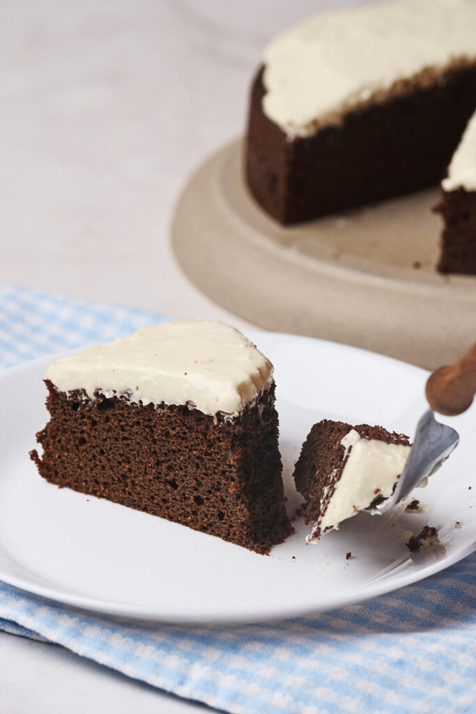 A slice of dark chocolate cake with white frosting on a white plate, with a fork lifting a small piece.