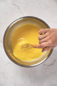 A hand whisking a yellow mixture in a stainless steel bowl on a white marble surface.