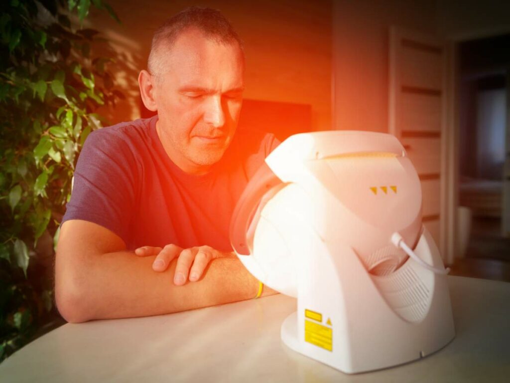 A man in a blue shirt sits at a table, using a white light therapy device emitting an orange glow, with plants and a partially opened door in the background.