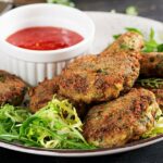 A plate with several crispy vegetable patties garnished with greens, accompanied by a small dish of red dipping sauce.