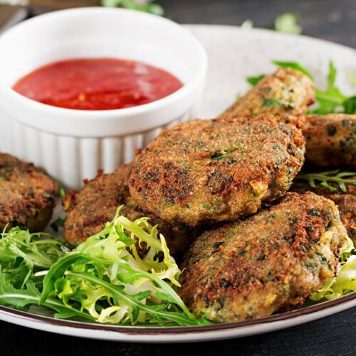 A plate with several crispy vegetable patties garnished with greens, accompanied by a small dish of red dipping sauce.