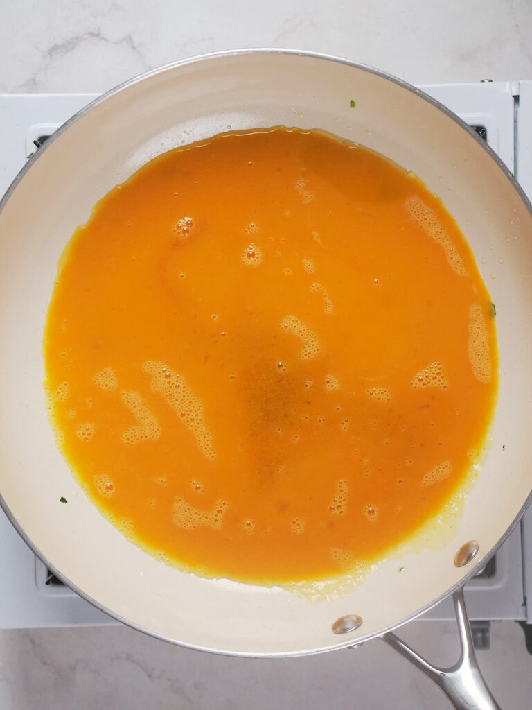 A frying pan on a stove contains beaten eggs being cooked, with visible bubbles forming on the surface.