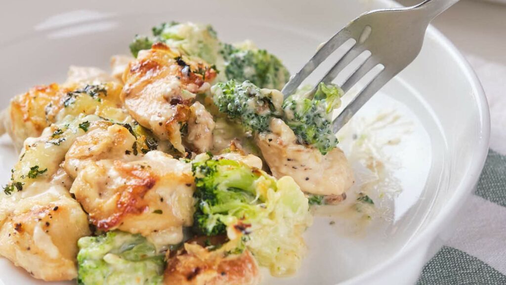 A fork lifts a piece of broccoli from a dish of cheesy broccoli and chicken casserole on a white plate.