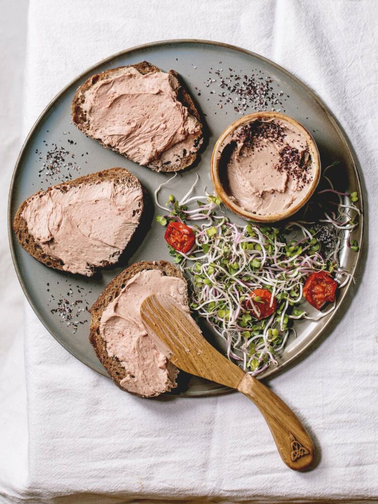 Plate with three slices of bread topped with pâté, a bowl of pâté, a wooden spreader, and a side salad of sprouts and tomatoes.