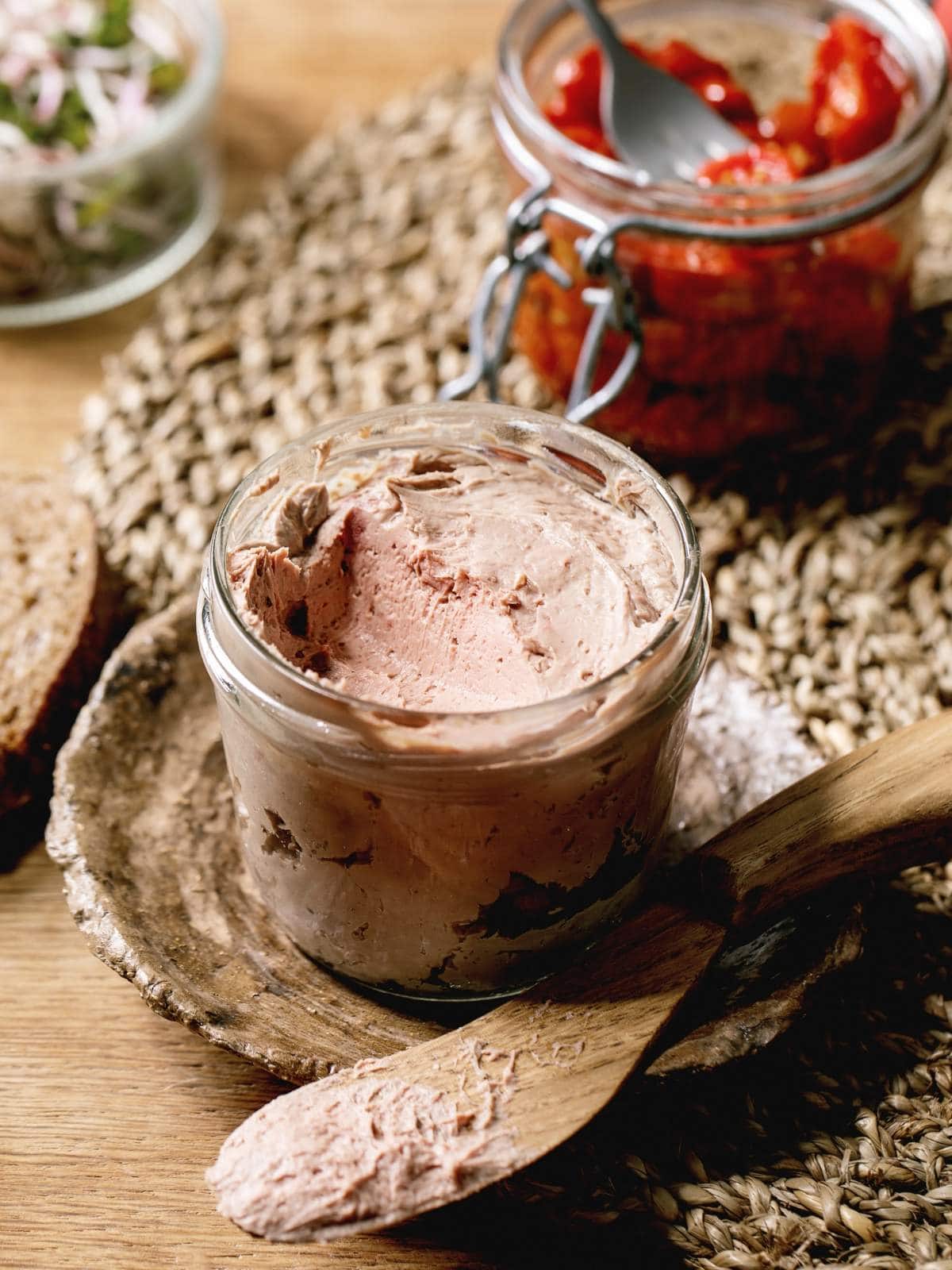 A glass jar of pâté with a wooden butter knife resting on a woven mat beside it.