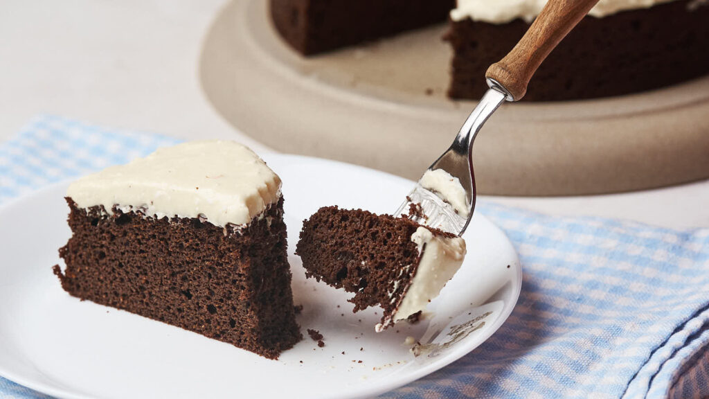 A fork lifts a piece of chocolate cake, topped with white frosting, from a plate.