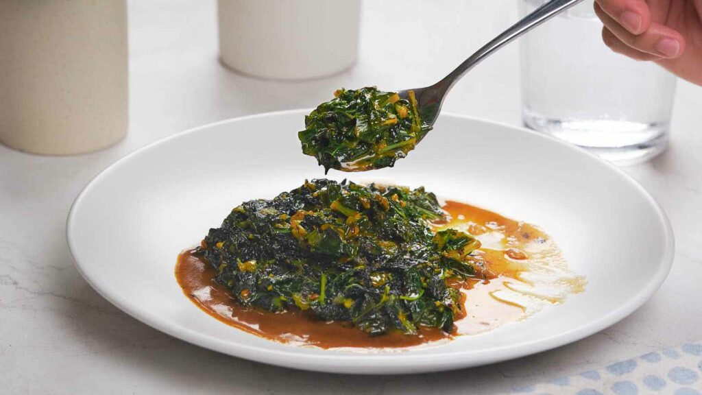 A person holding a spoonful of cooked leafy greens over a white plate containing more greens in a sauce. The background has two white cups and a clear glass of water.