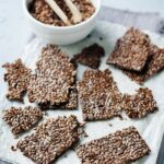 A bowl of flaxseeds with a wooden scoop next to pieces of flaxseed crackers on a white cloth.