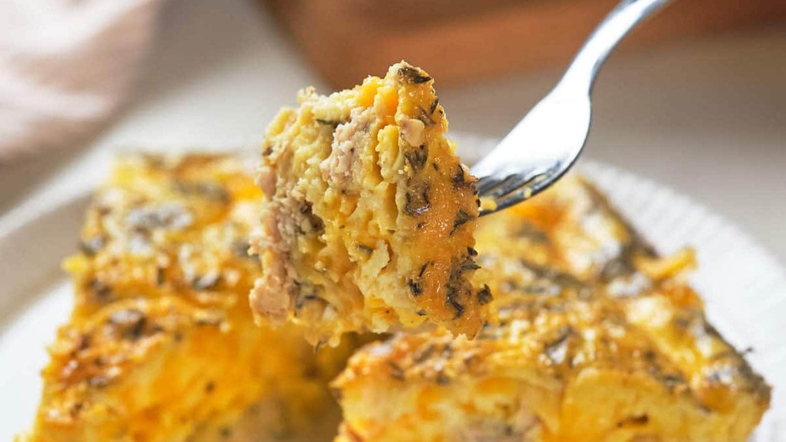 A fork holding up a bite of a savory casserole with visible herbs and cheese, against the backdrop of a plate with two square slices of the casserole.