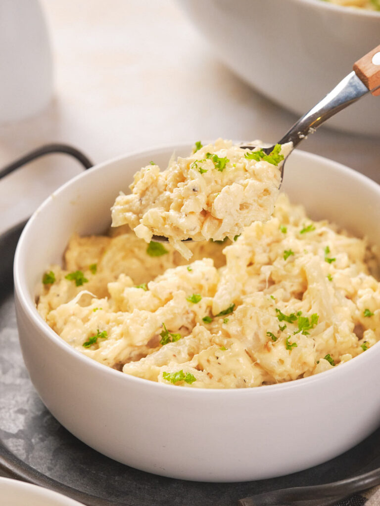 A bowl of creamy casserole garnished with green herbs, with a spoon lifting a portion.