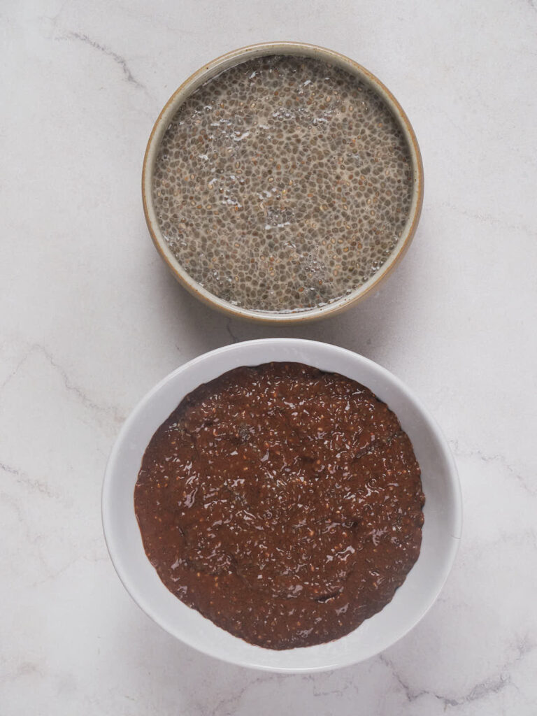 Two bowls on a marble surface, the top contains a gray chia seed pudding and the bottom holds a thick reddish-brown sauce or spread.