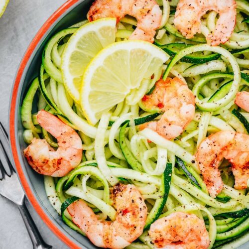 A bowl of zucchini noodles topped with grilled shrimp and lemon slices, with a fork and knife on the side.
