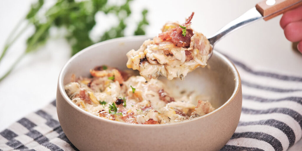 A spoon scooping creamy chicken casserole with bacon and parsley from a bowl placed on a striped cloth.