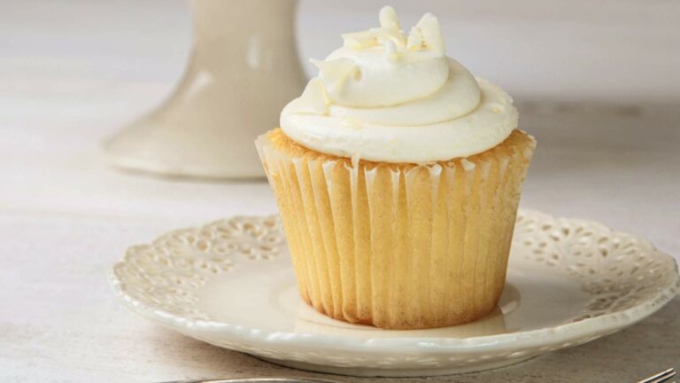 A vanilla cupcake with white frosting and white chocolate shavings sits on a decorative plate.