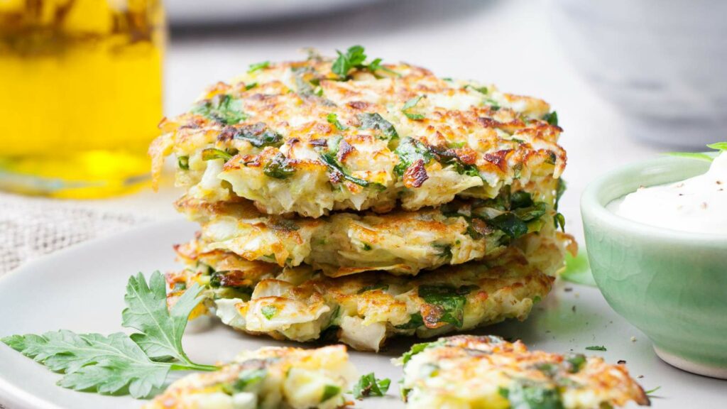 Three cabbage fritters stacked on a plate with a bowl of yogurt sauce on the right and a sprig of parsley on the left.