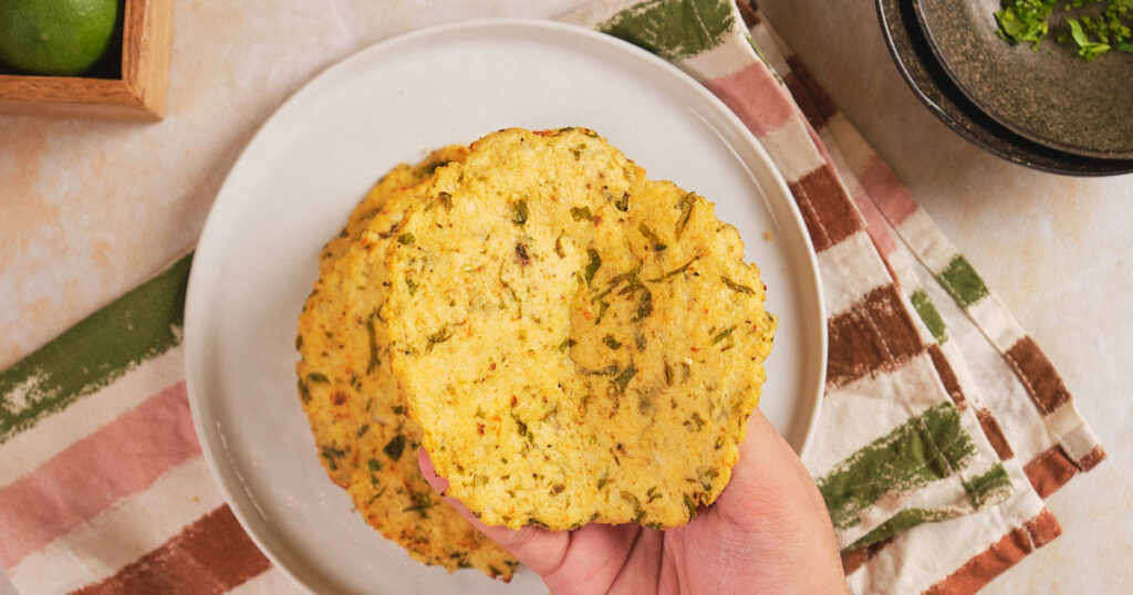A hand is holding several gluten-free tortillas with visible herbs on a white plate, placed on a striped cloth.