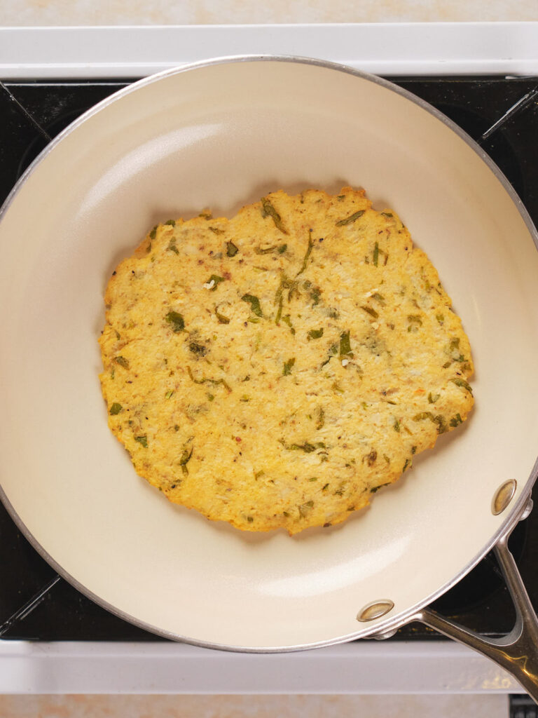 A large, round, cauliflower tortilla with green herb flecks cooking in a white frying pan on a stovetop.