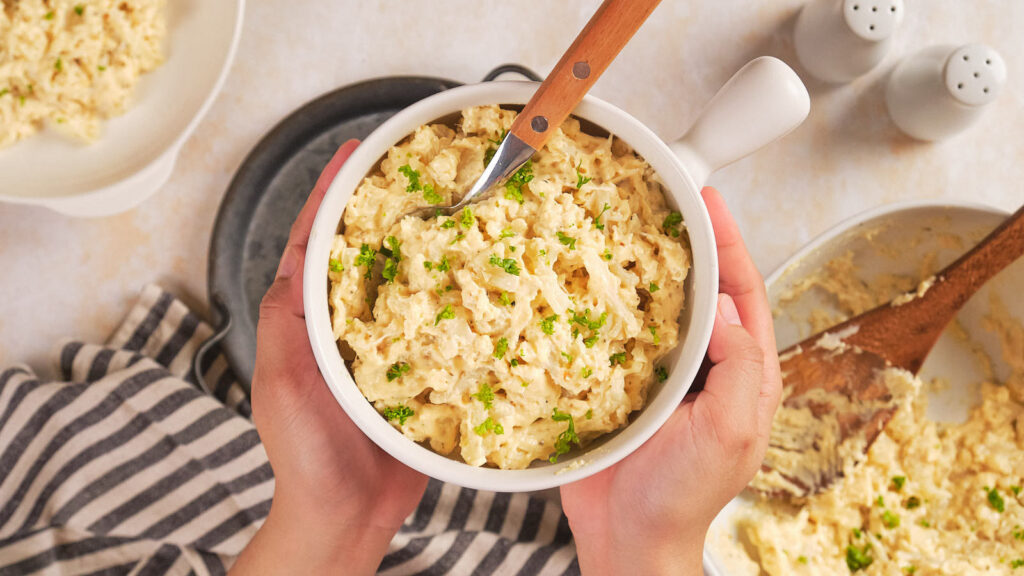 Hands holding a white bowl of macaroni salad garnished with chopped herbs.