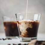 Cream being poured into cold brew black coffee in a glass, creating a swirl effect.