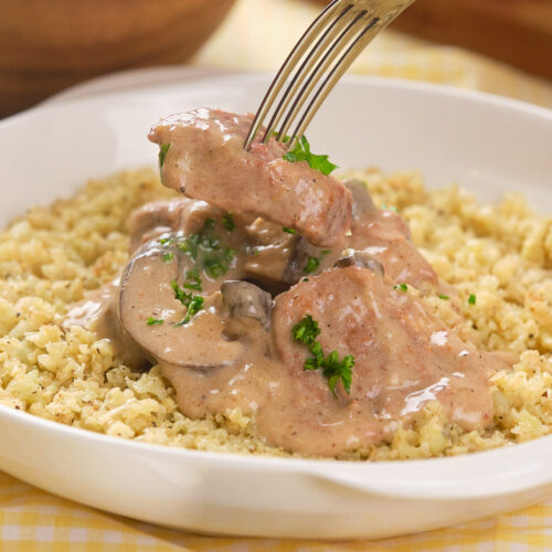 A fork lifts a piece of beef over a plate of creamy slow cooker stroganoff on a white dish.