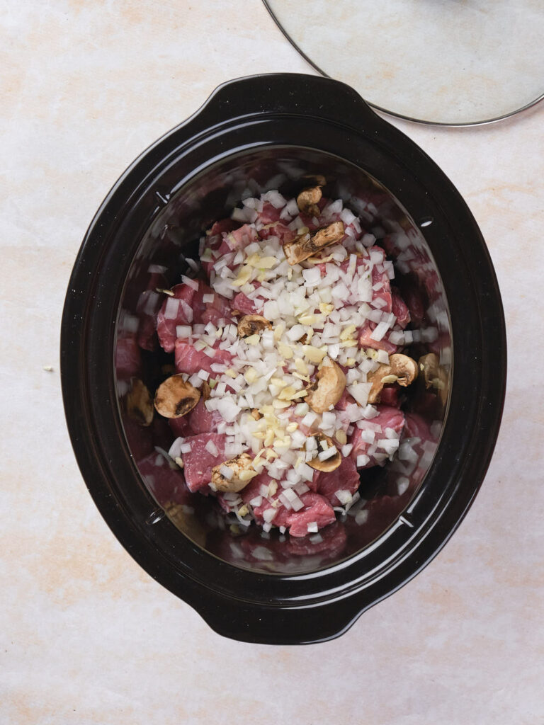 A slow cooker filled with raw cubed beef, chopped onions, garlic, and mushrooms.