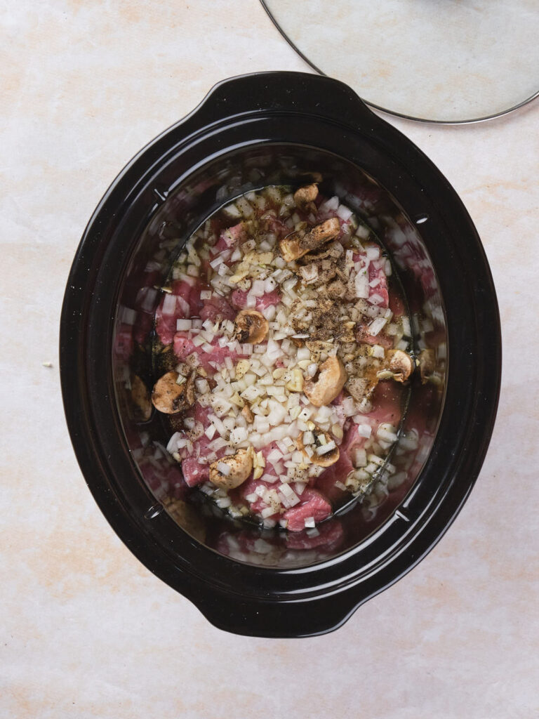 Top view of a slow cooker filled with raw diced meat, chopped onions, mushrooms, and seasoning.