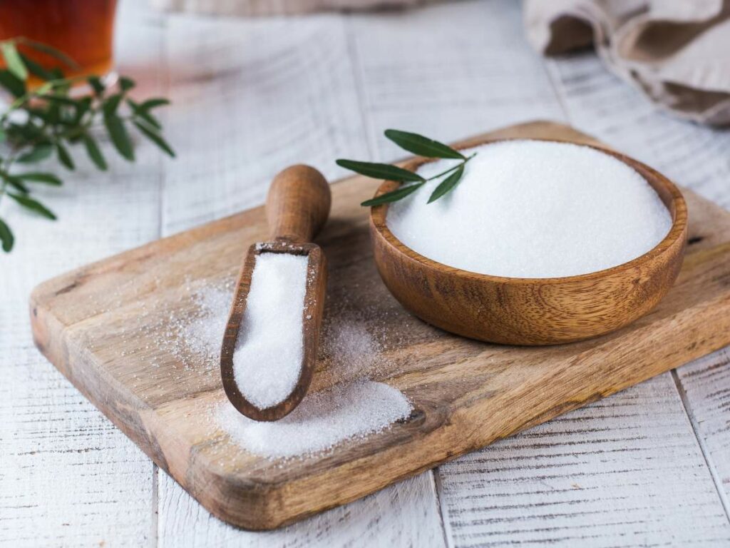 A wooden bowl and scoop filled with white sugar on a wooden cutting board, with a green sprig nearby.