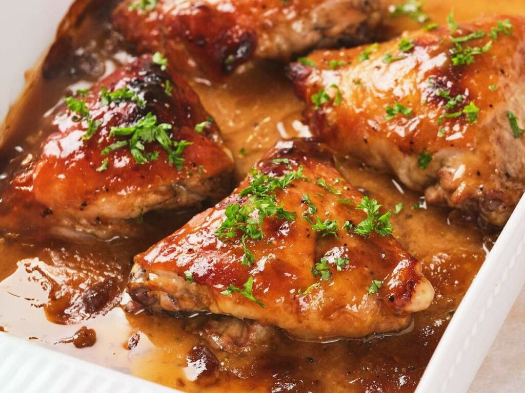 Baked chicken thighs in apricot sauce, garnished with chopped parsley, in a white baking dish.