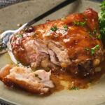 A piece of aptricot glazed cooked chicken partially sliced on a plate, garnished with herbs, with a fork beside it.