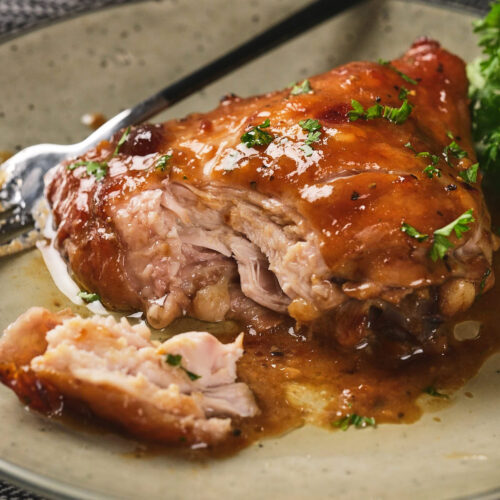 A piece of aptricot glazed cooked chicken partially sliced on a plate, garnished with herbs, with a fork beside it.