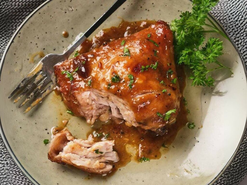 A glazed piece of cooked meat with herbs on a plate, accompanied by a fork and garnished with parsley.