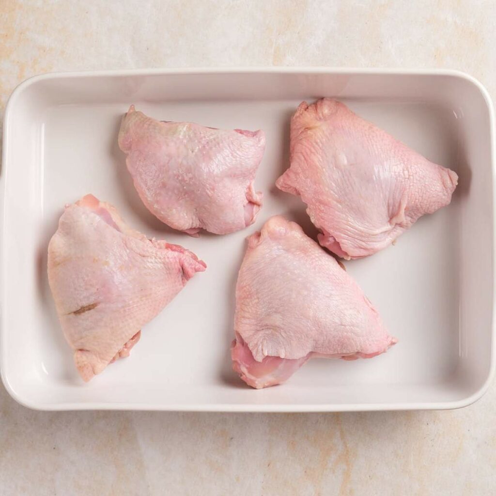 Four raw chicken thighs in a white rectangular dish on a light surface.