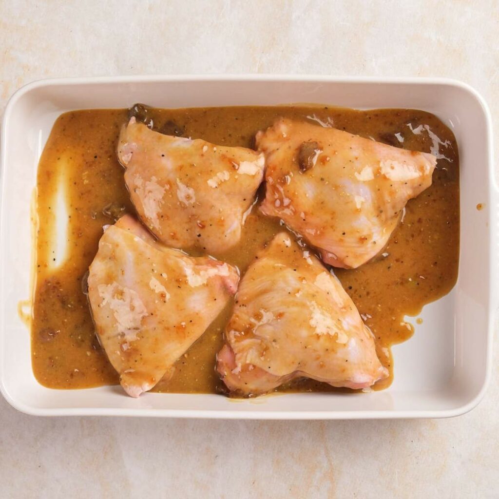 Four raw chicken thighs in a white baking dish, coated with a thick brown marinade.