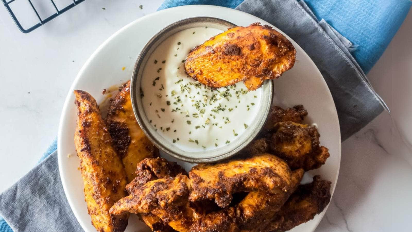 Plate of blackened chicken tenders with a bowl of creamy dipping sauce, garnished with herbs.