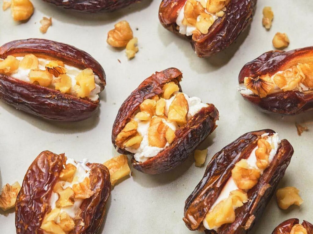 Dates stuffed with cream cheese and topped with chopped nuts on a white surface.