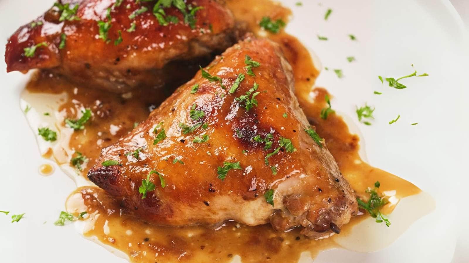 Two apricot chicken thighs on a plate, topped with chopped herbs and surrounded by a light brown sauce.