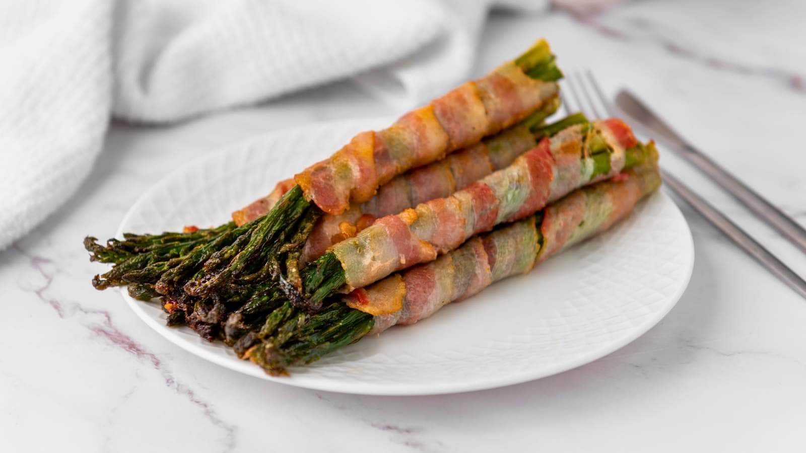 A plate of bacon wrapped asparagus, neatly arranged in a row, with a fork and knife beside it on a marble surface.