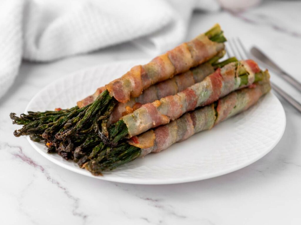 Asparagus stalks wrapped in crispy bacon, arranged on a white plate with a marble countertop background.