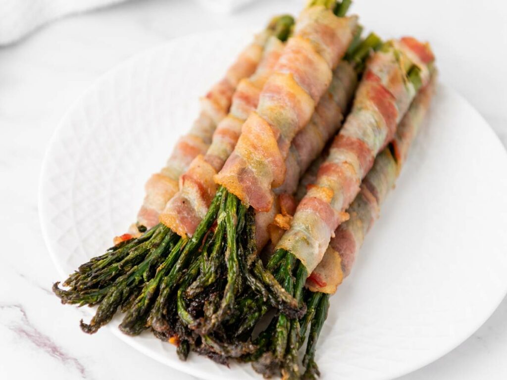 Bundles of asparagus wrapped in cooked bacon are neatly arranged on a white plate.