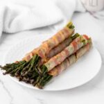 Bacon-wrapped asparagus spears arranged on a white plate atop a marble surface.