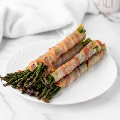 Bacon-wrapped asparagus spears arranged on a white plate atop a marble surface.