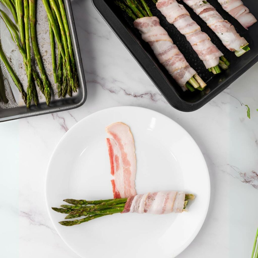 Bacon-wrapped asparagus on a white plate, next to trays of plain and wrapped asparagus, placed on a marble surface.