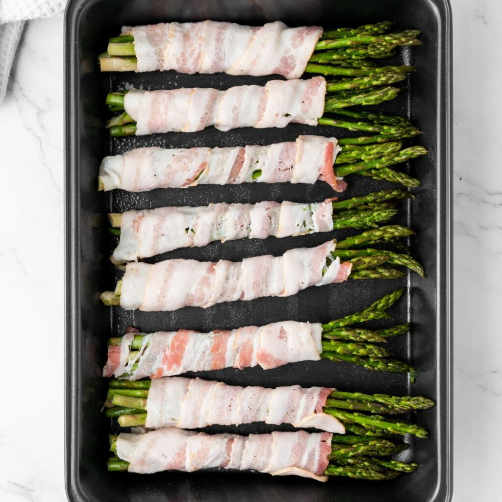 Bacon-wrapped asparagus spears arranged in a baking tray on a marble surface.