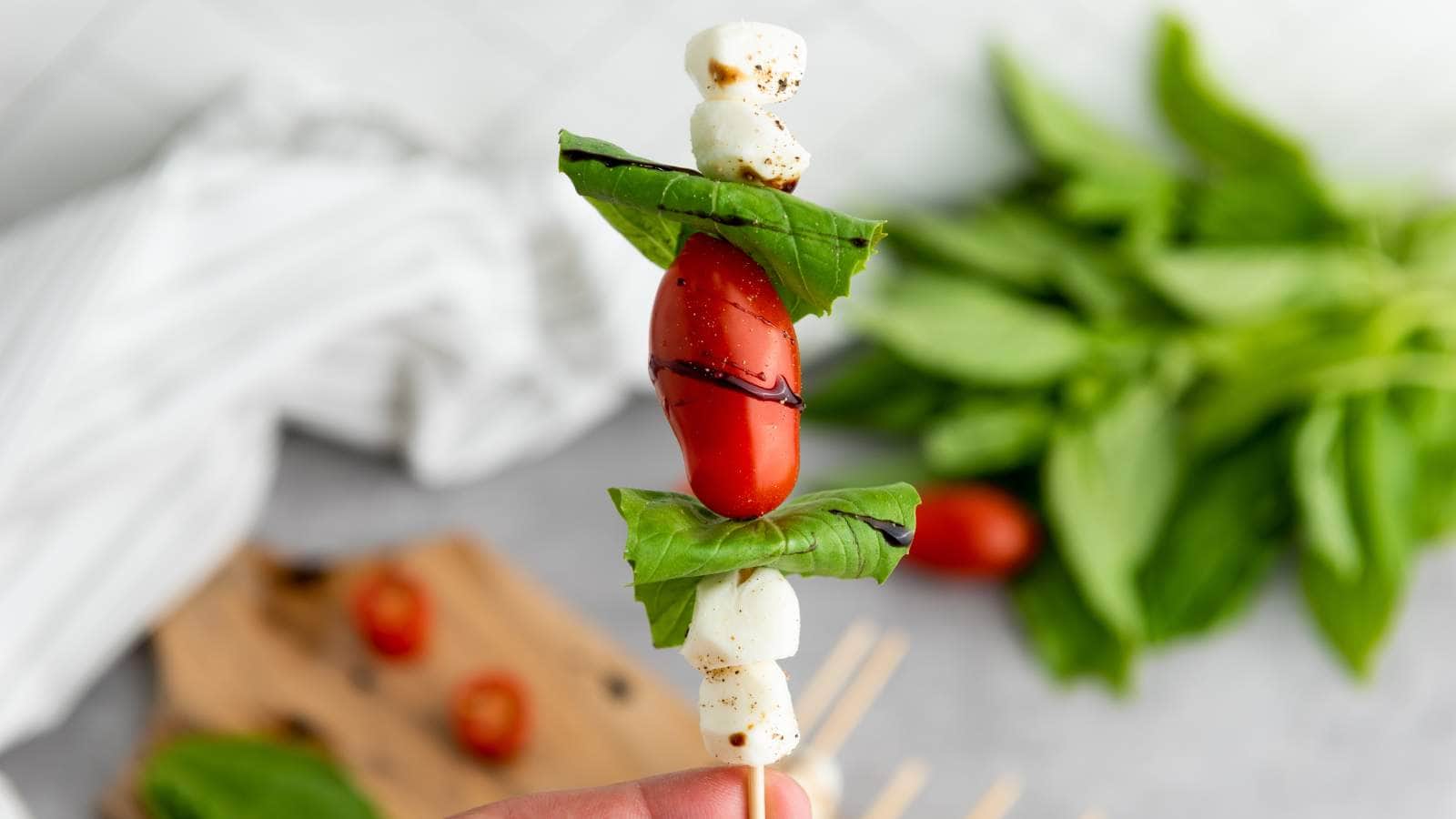 A caprese appetizer with basil leaves, a cherry tomato, and mozzarella balls, drizzled with balsamic glaze.